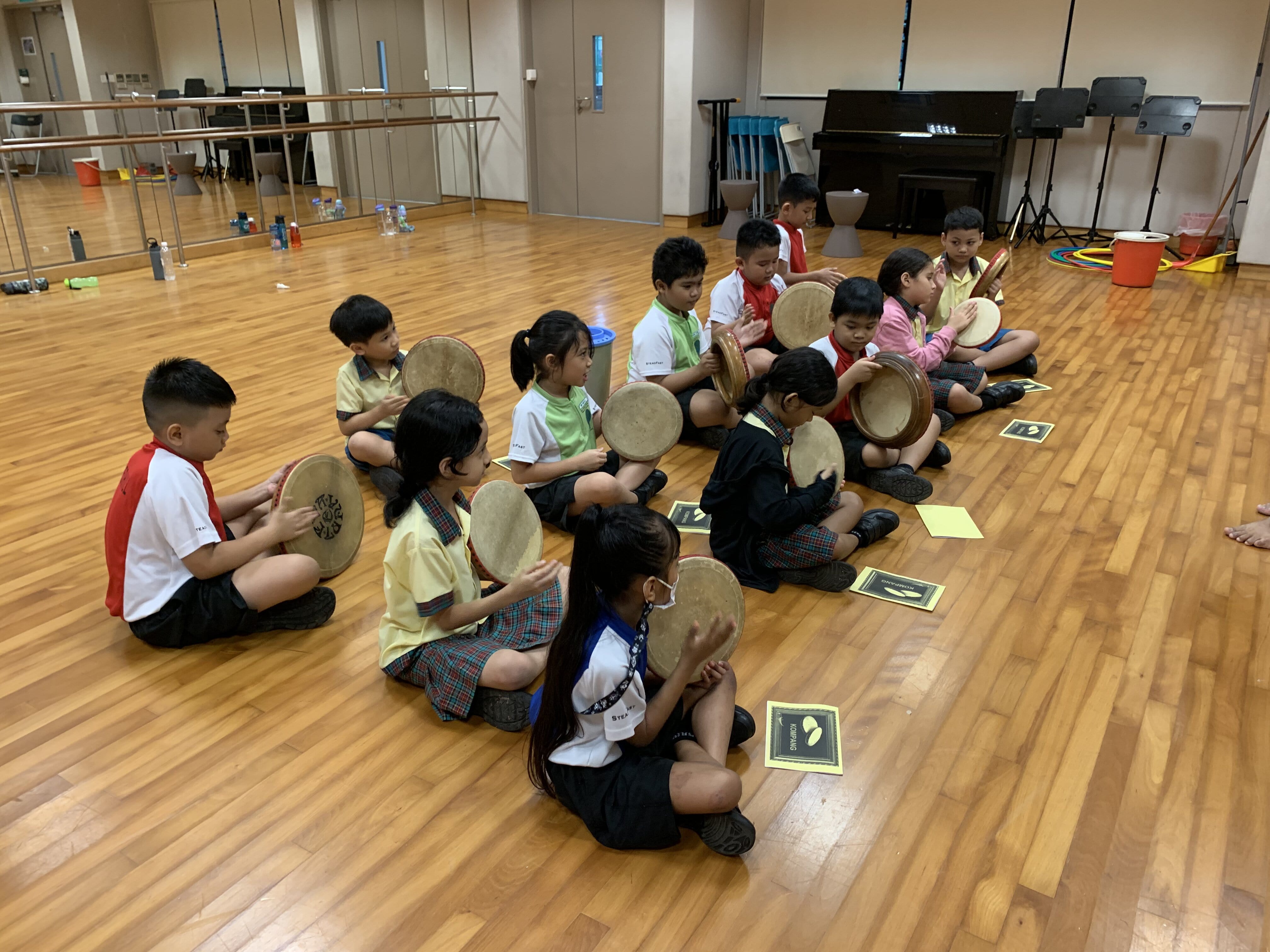 Children in school uniforms sit on a wooden floor in a circle, each holding and playing a round hand drum.